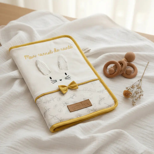 Protège carnet de santé bébé lapin blanc coton bio avec oreilles 3D, broderie dorée et nœud moutarde, posé sur mousseline blanche avec hochet bois - cadeau naissance original